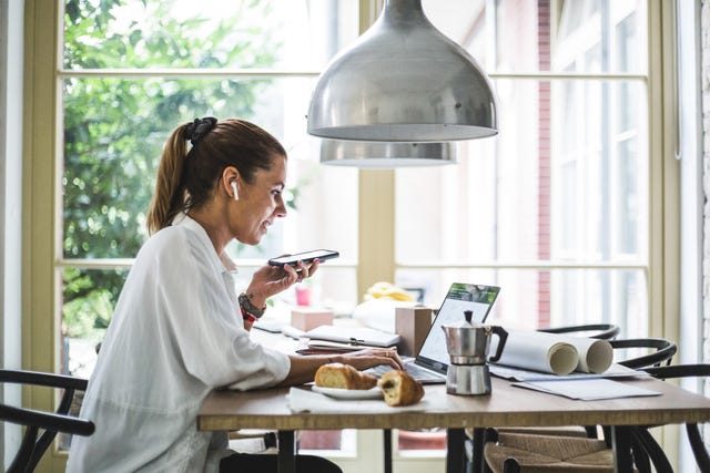 smiling female entrepreneur using laptop while talking through smart phone at home office