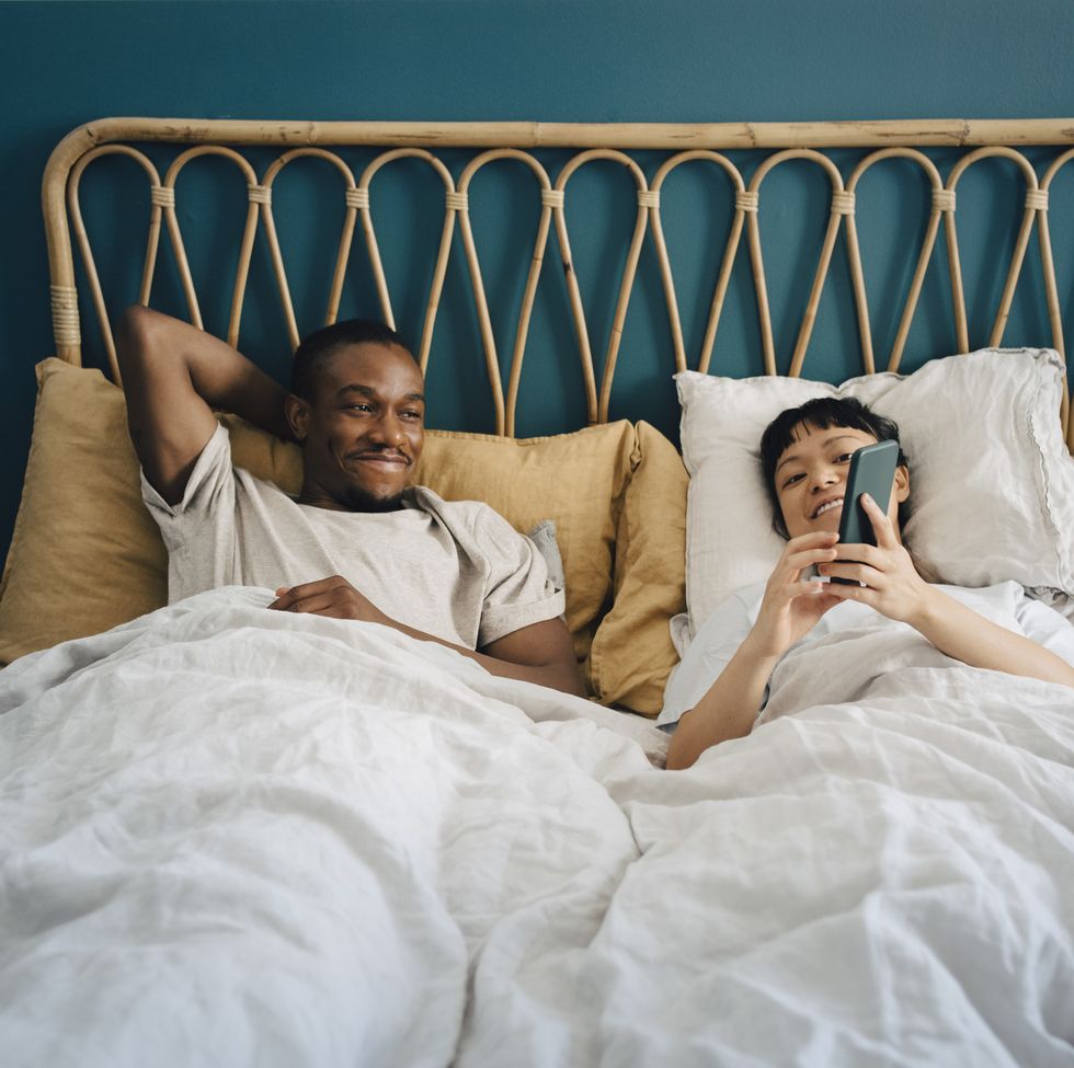 smiling girlfriend sharing smart phone with boyfriend while lying on bed
