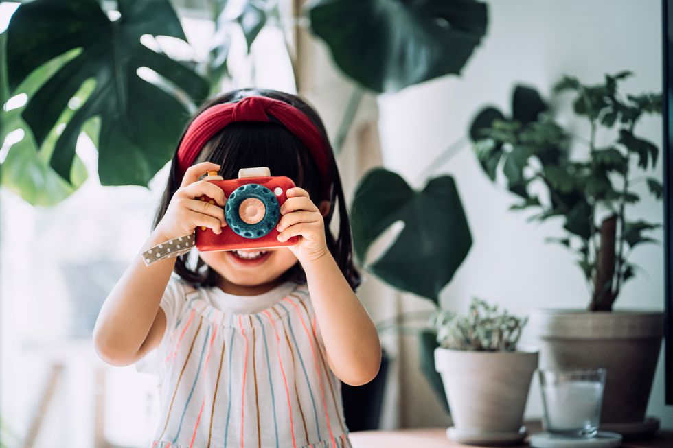 smiling little asian girl with red headband acting like a professional photographer having fun while taking photos with wooden toy camera in front of potted plants at home