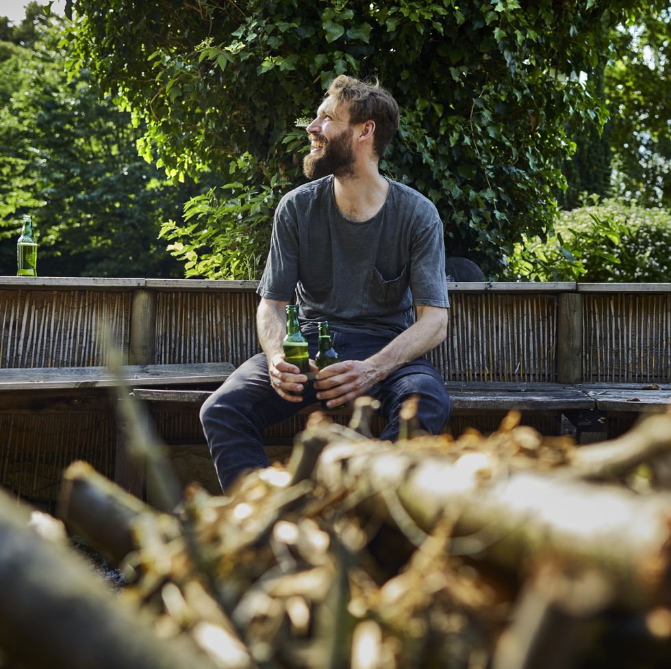 Smiling man holding beer bottles sitting on bench in garden