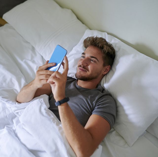 smiling man using smart phone while lying on bed in bedroom