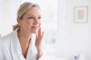 smiling mature woman applying moisturizer to face at bathroom mirror