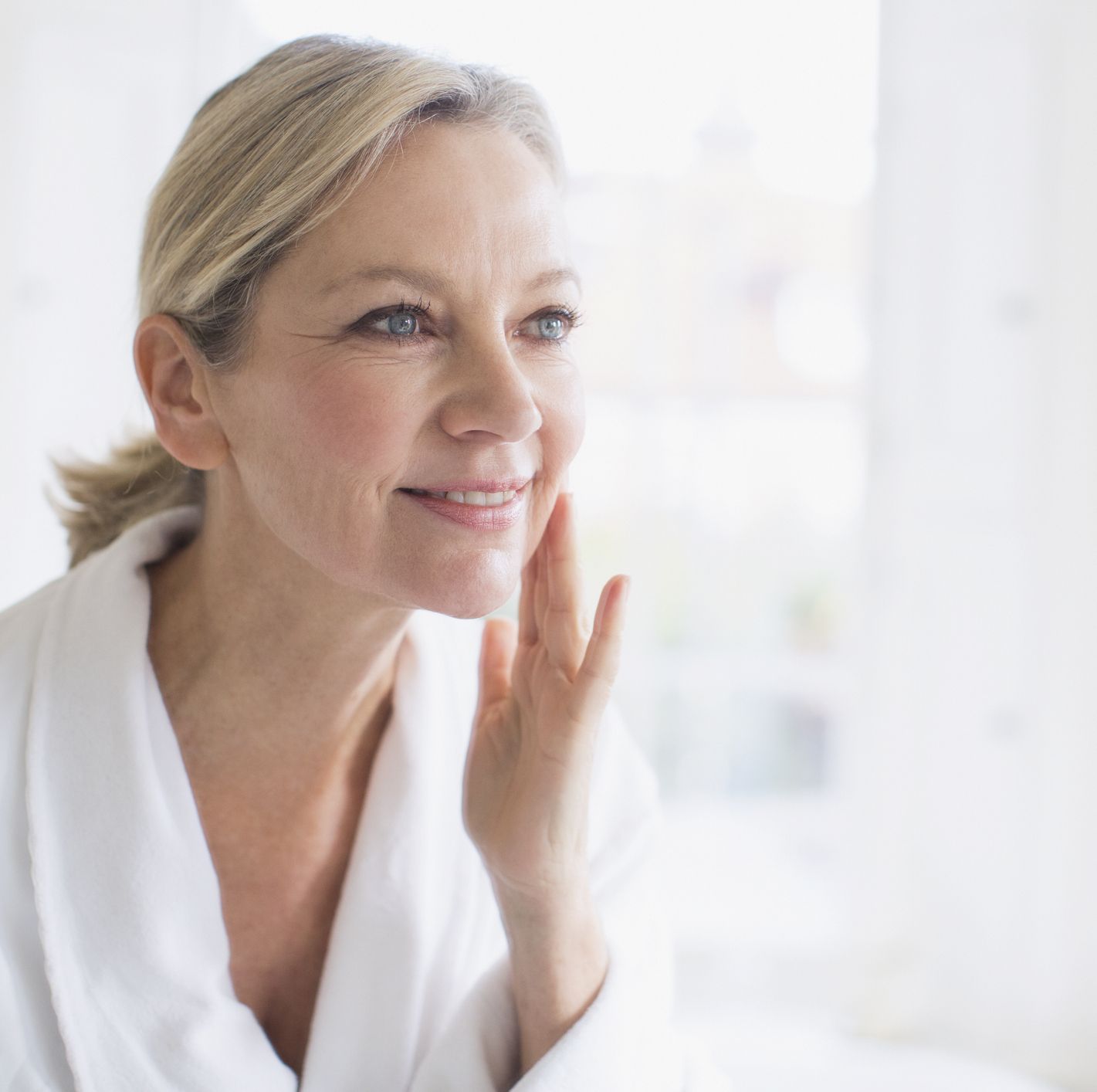 Smiling mature woman applying moisturizer to face at bathroom mirror