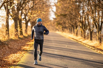 smiling middle aged man dressed sporty in black leggings and blue jacket running through forest full of fallen golden leaves on beautiful autumn day