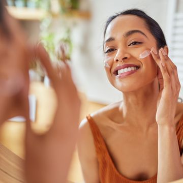 smiling mixed race young woman applying moisturizer on her face in bathroom