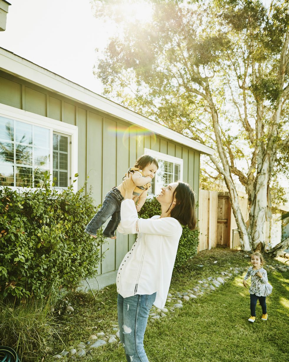 smiling mother holding infant son in air in front yard of home on summer morning