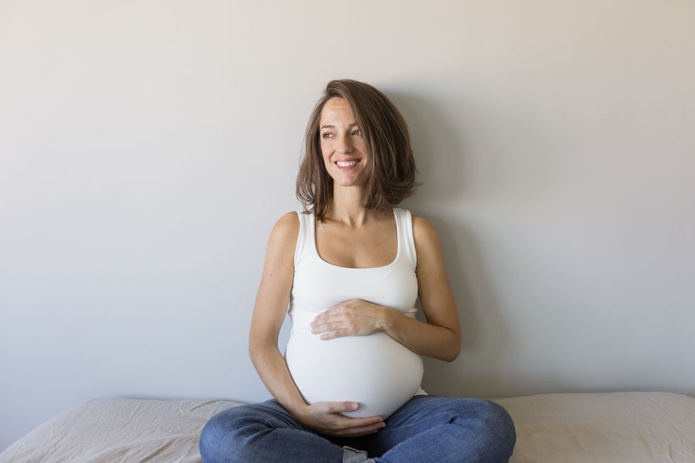 smiling pregnant woman touching belly while sitting on bed at home