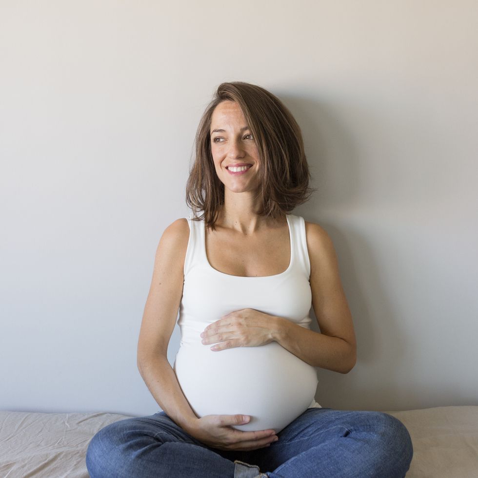 smiling pregnant woman touching belly while sitting on bed at home