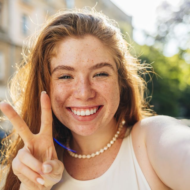 smiling redhead woman showing peace gesture while taking selfie