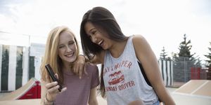 Smiling teenage girls with skateboard texting with cell phone in skate park