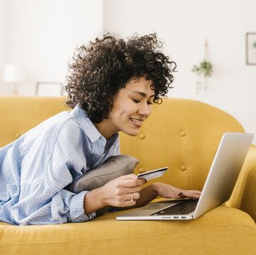 smiling woman holding credit card using laptop lying on sofa in living room at home