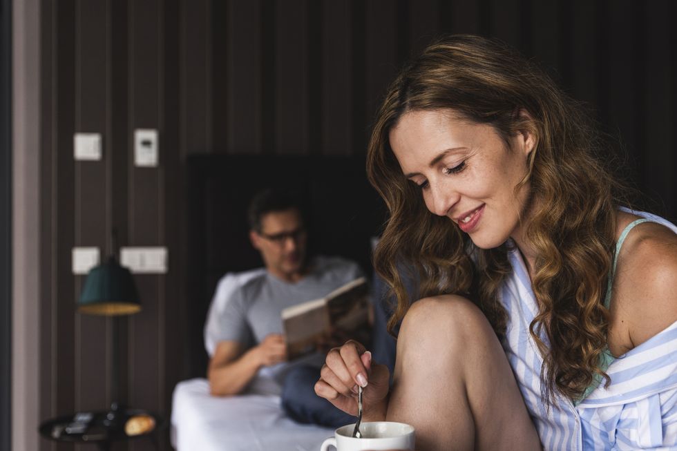 Smiling woman with cup of coffee in bedroom with man in background