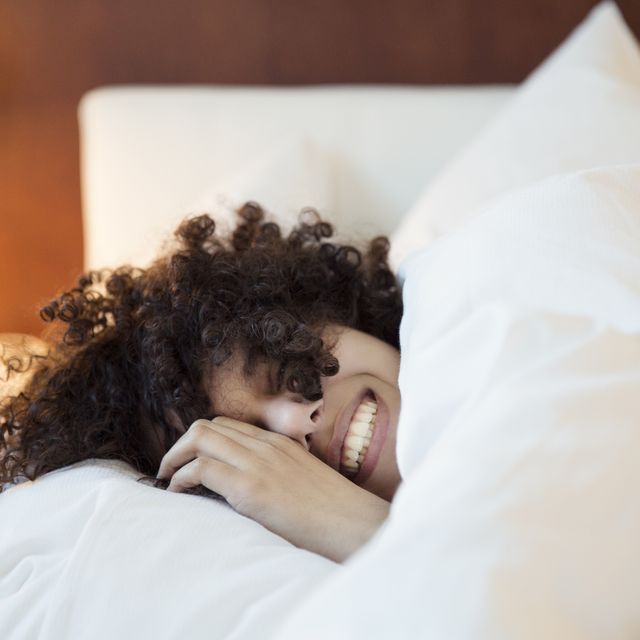 a smiling woman with dark curly hair in bed under the covers, head on a white pillow,