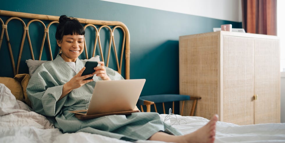 smiling woman with laptop using smart phone on bed at home