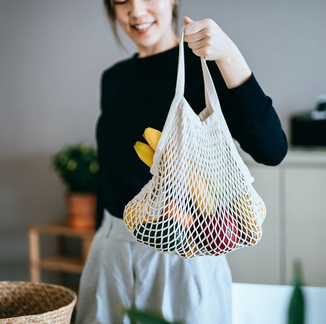 smiling young asian woman coming home from grocery shopping she is holding a reusable mesh bag full of fresh and healthy organic fruits and vegetables responsible shopping, zero waste, sustainable lifestyle concept