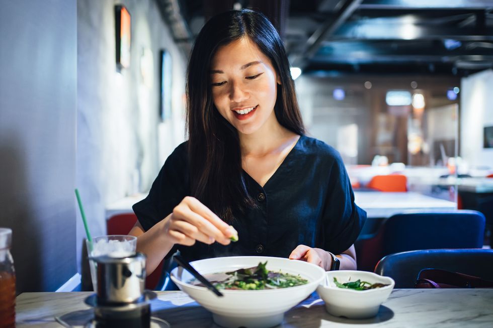 smiling young asian woman enjoying meal in a restaurant she is squeezing fresh lime on her freshly served traditional vietnamese pho soup with noodles and beef, with vietnamese iced coffee by the side