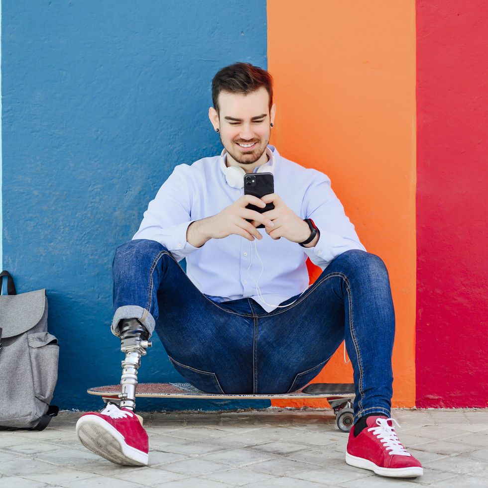 smiling young man with leg prosthesis and skateboard using smartphone
