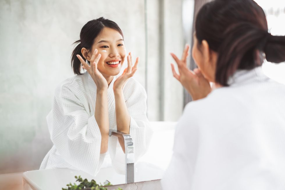 smiling young woman applying moisturizer on face on mirror in bathroom