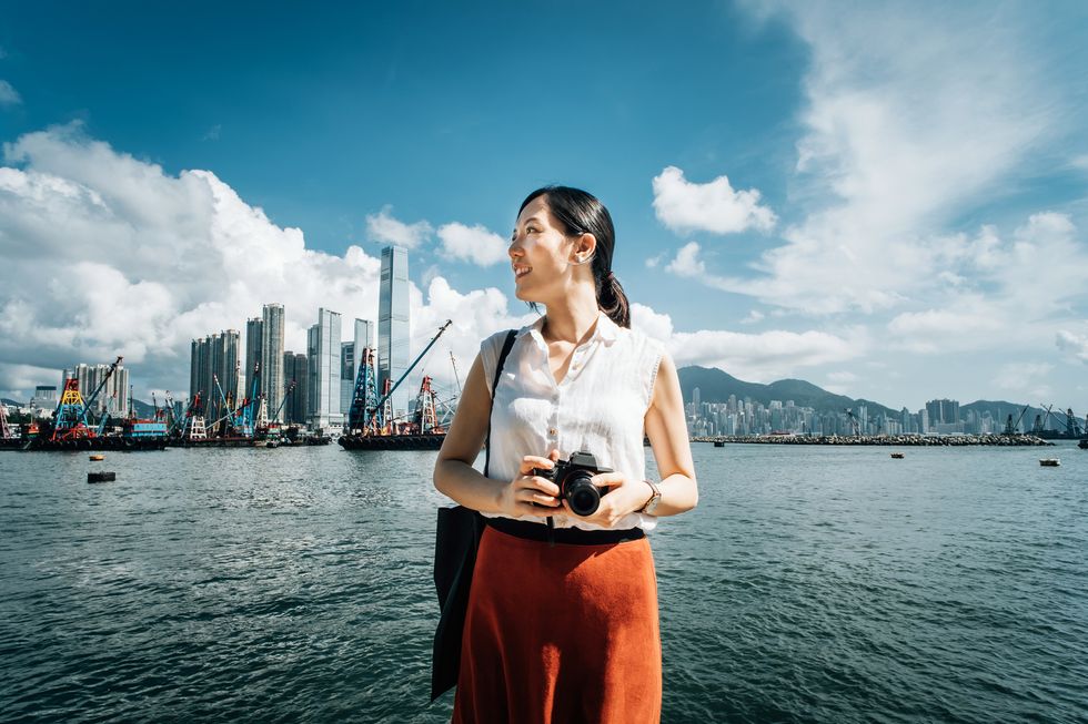 smiling young woman photographing with camera against iconic city skyline and busy commercial dock on a sunny day