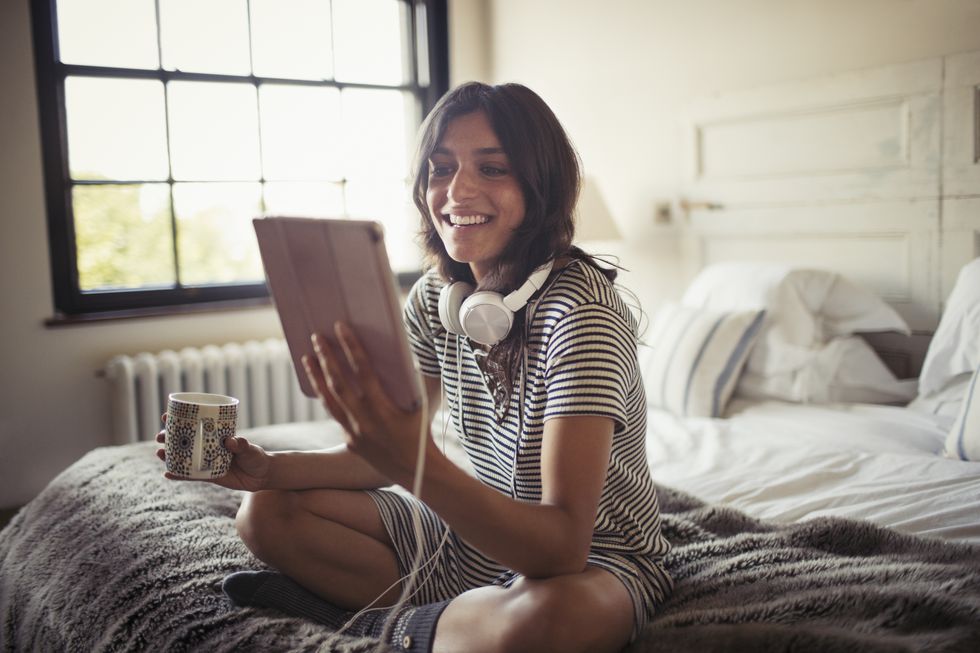 Smiling young woman with headphones drinking coffee and using digital tablet on bed