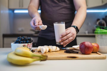 man preparing a protein drink in a kitchen, holding a portion of whey protein powder in a measuring spoon and a glass of milkshake or smoothie in his hand.