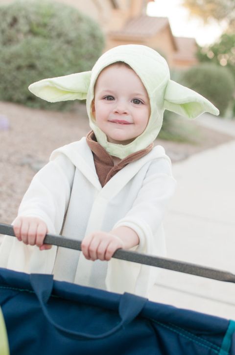 toddler in yoda costume, with white robe and light green head piece with big ears, holding on to cart