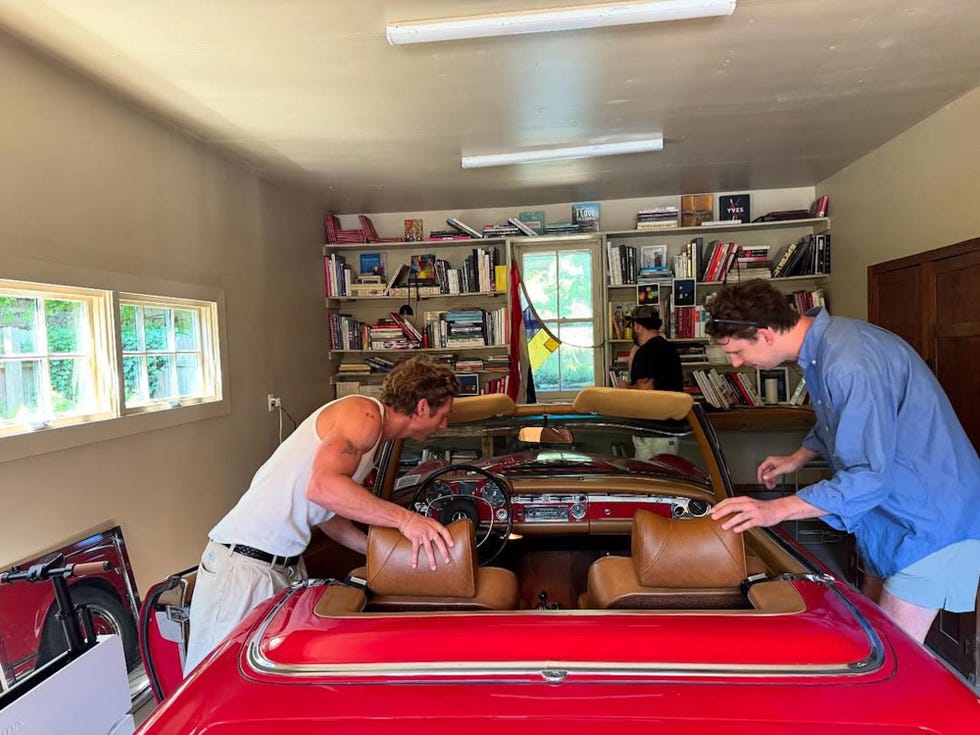 Two individuals riding in a classic red convertible car in a garage