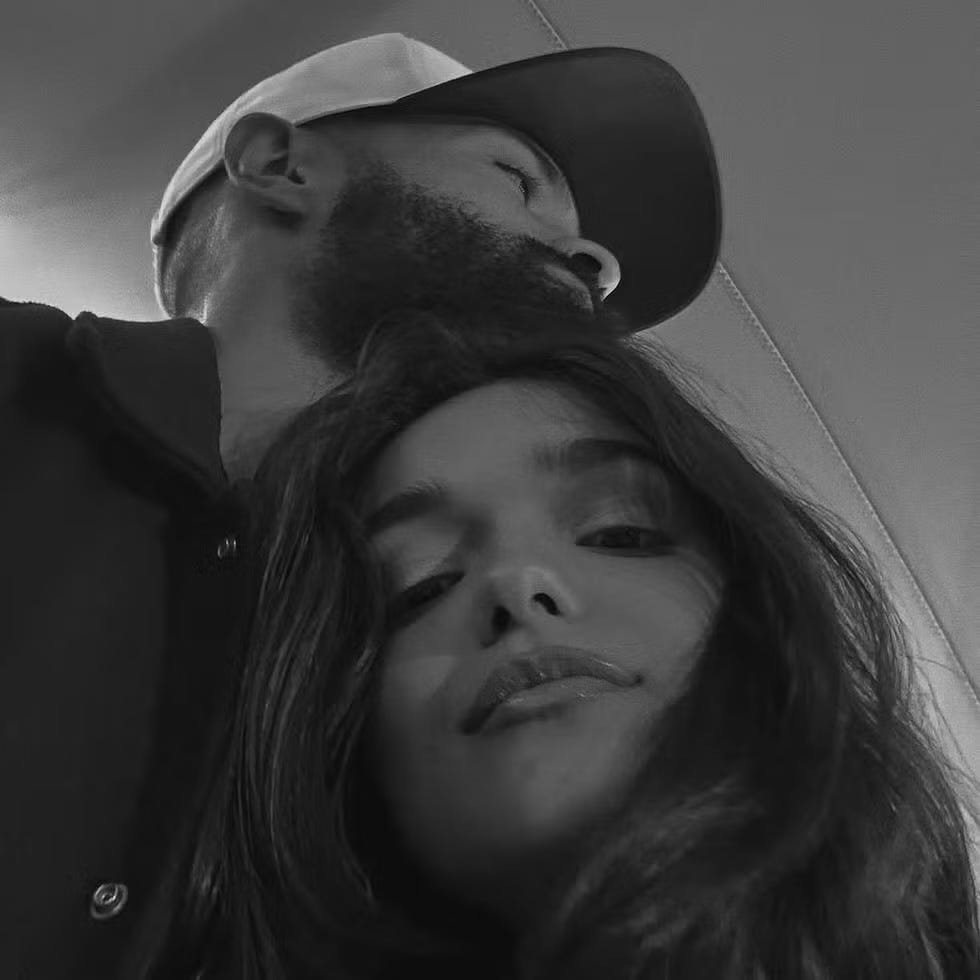 a bearded man wearing a cap is seen leaning over a person with long hair