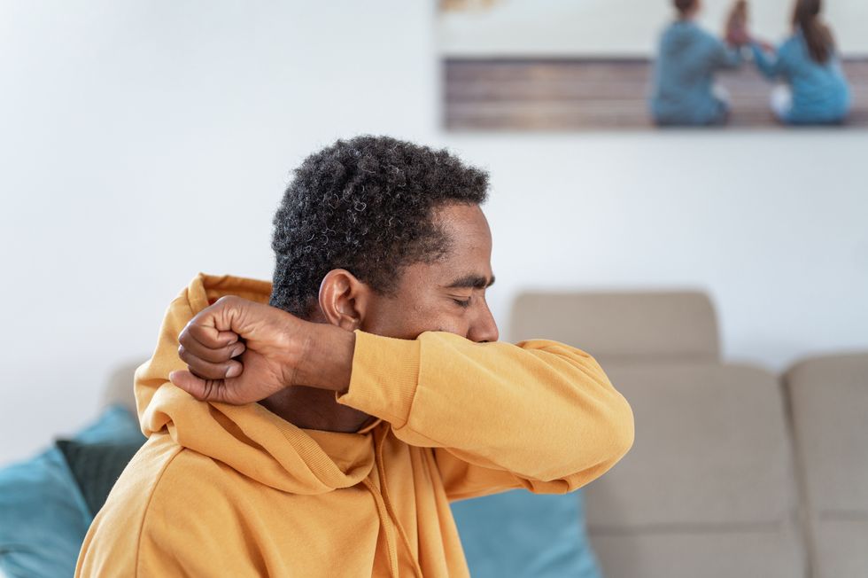 adult man wearing a yellow hoodie in a living room, coughing or sneezing into elbow