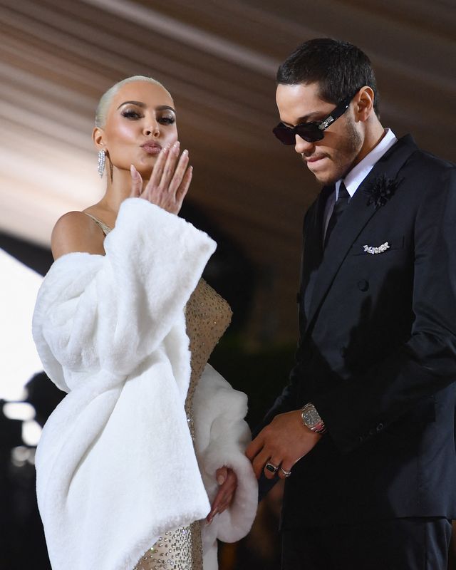 kim kardashian, wearing a white fur coat and white sequined dress, blows a kiss at the camera while pete davidson, wearing a black suit and sunglasses, stands next to her and looks down