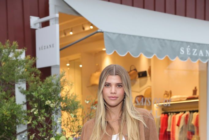 sofia richie poses for a photo while standing in front of a store, she wears a brown jacket over a white top and black jeans