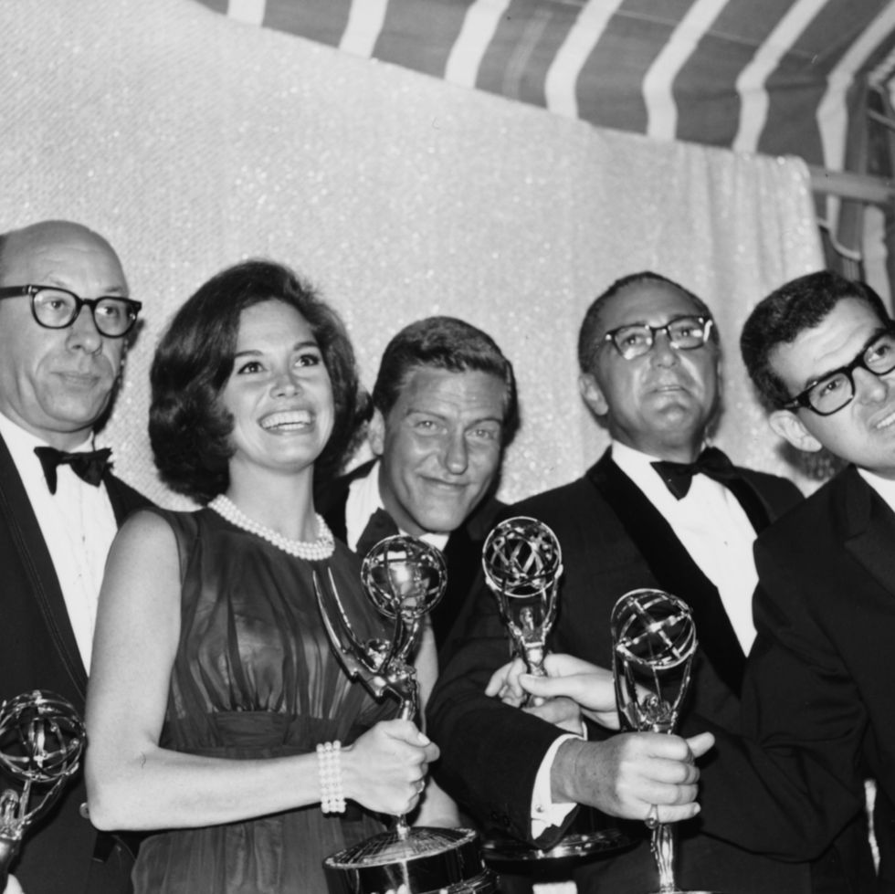 richard deacon, mary tyler moore, dick van dyke, and sheldon leonard stand while holding their emmy statuettes, the men are wearing tuxedos and a smiling moore wears a dress