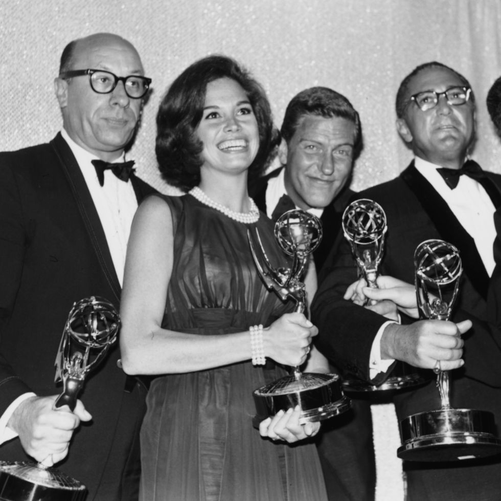 richard deacon, mary tyler moore, dick van dyke, and sheldon leonard stand while holding their emmy statuettes, the men are wearing tuxedos and a smiling moore wears a dress