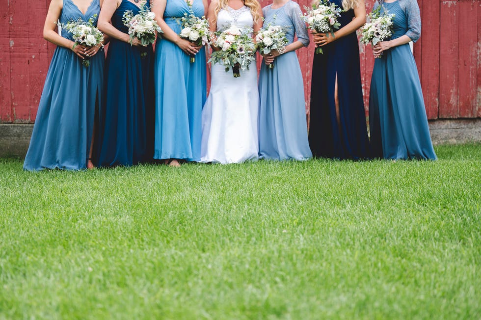 a bride in a white dress and bridesmaids in bridesmaids dresses in various shades of blue standing with bouquets on a lawn