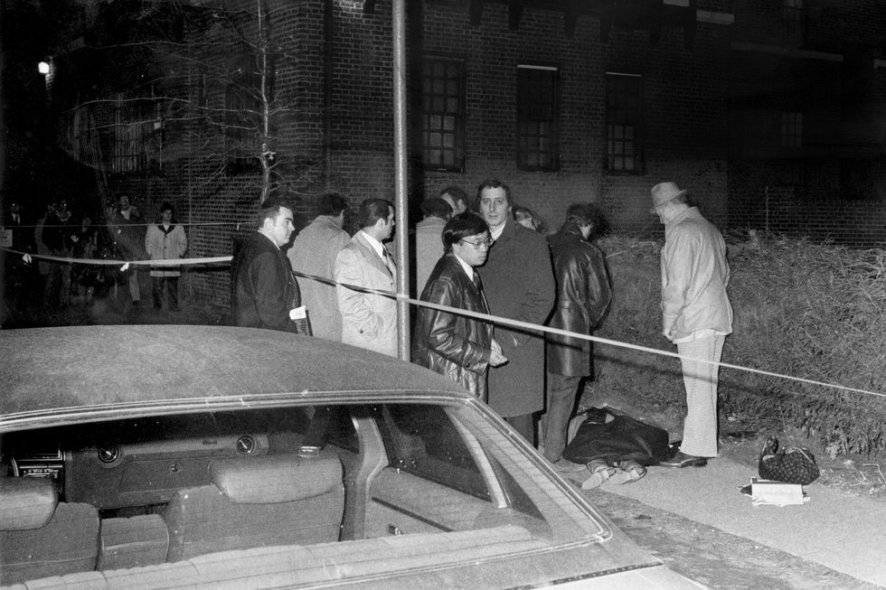 a group of men stand around a sidewalk as a person lies on the ground, a car and police tape appear in the foreground