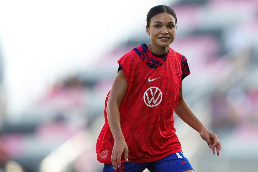 sophia smith looks at the camera while moving to the right, she wears a red and blue usa soccer uniform with a red penny over top that has nike and vw logos