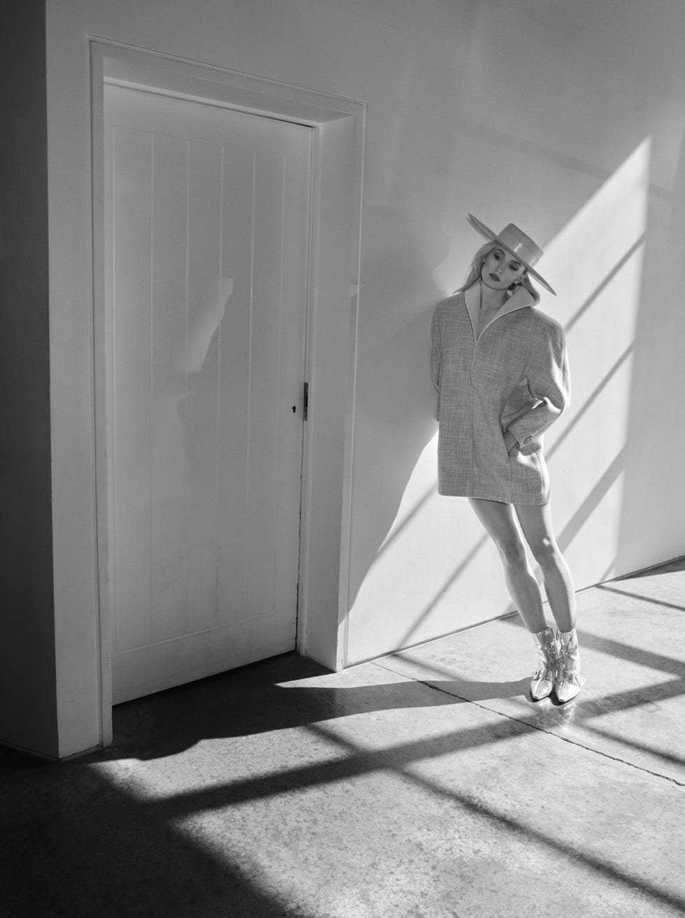 a figure stands against a wall wearing an oversized coat with a high collar complemented by shiny silver boots