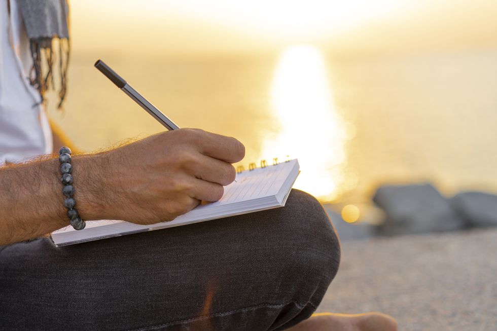 man writing on a notebook during sunrise on rocks at the beach