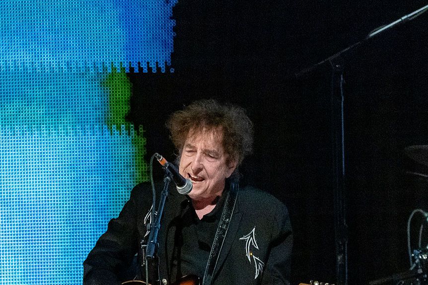bob dylan sings into a microphone and plays electric guitar while seated on a stool