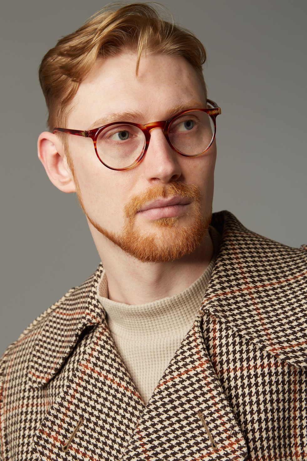 a man with ginger hair and a beard wearing tortoise shell glasses and a houndstooth coat