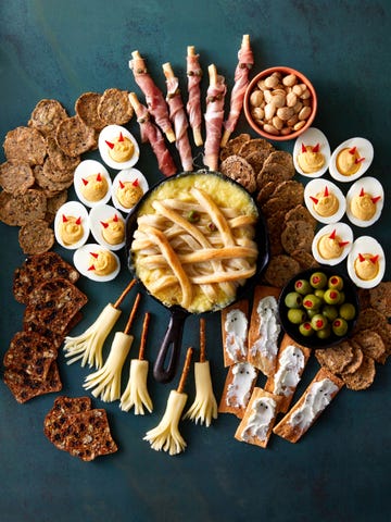 a spooky charcuterie board with olives, crackers, nuts, deviled eggs with little horns in em, crackers with smears of cheese that look like ghosts on em, broomsticks made of string cheese with pretzels stuck in em, and a baked brie that looks like a mummy