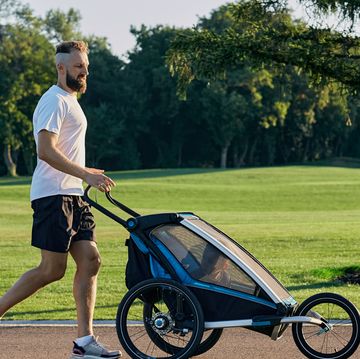 sporty man in sportswear running with baby stroller at sunny morning in city park during fatherhood time. side view
