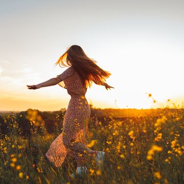 rear view of long haired woman in a dress dancing in a flower field raising arms at sunset