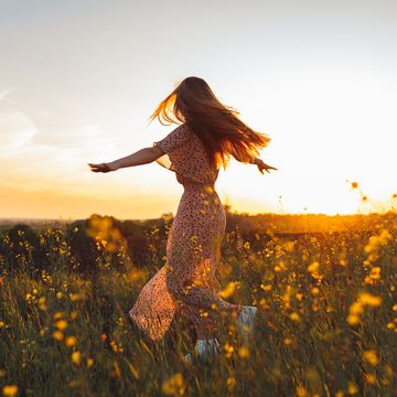 rear view of long haired woman in a dress dancing in a flower field raising arms at sunset