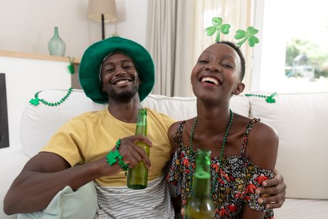 portrait of laughing african american couple in st patricks day costumes holding bottles of beer staying at home in isolation during quarantine lockdown