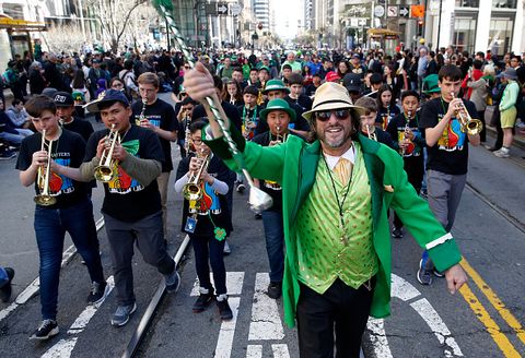 musicians march and perform with a band leader dressed in a straw fedora, green suit, light green vest and sunglasses leading them