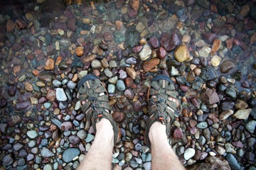 a topdown view of feet in water shoes standing on a lakeshore of colorful rocks