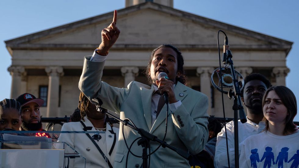 justin jones, wearing a gray suit and purple shit, stands in front of the tennessee state capitol on a stage with other people, speaking into a microphone and raising a finger in the air