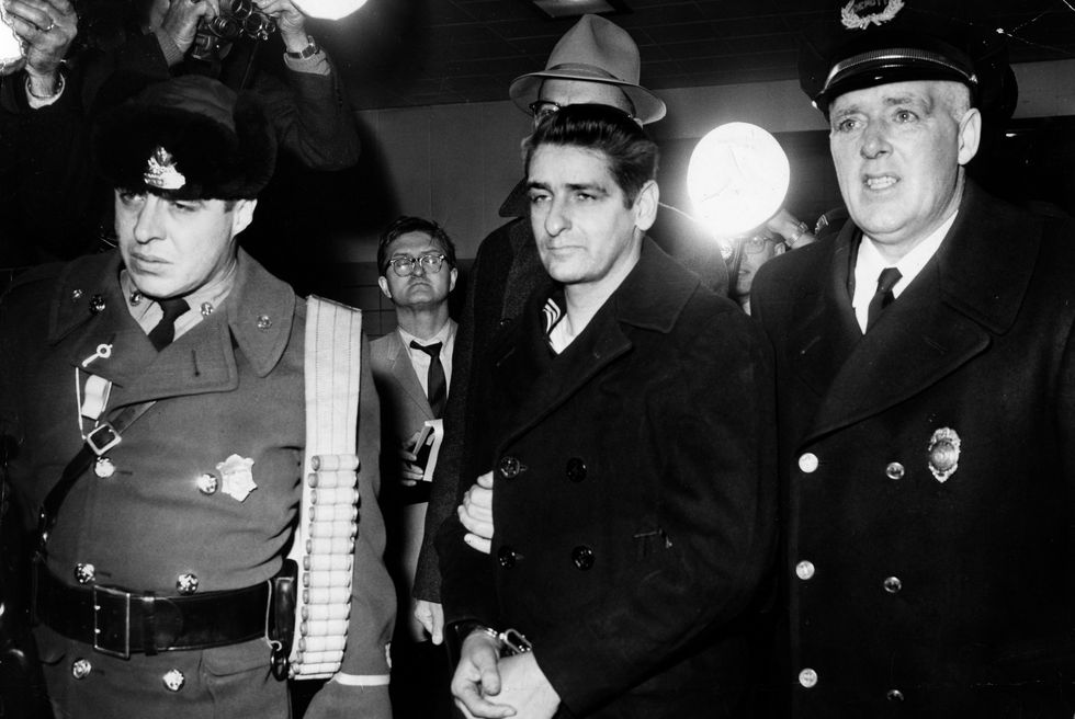 albert desalvo, wearing handcuffs and a dark peacoat, is led to prison by authorities, an officer is on either side of him, in the background are photographers, photography equipment and other men