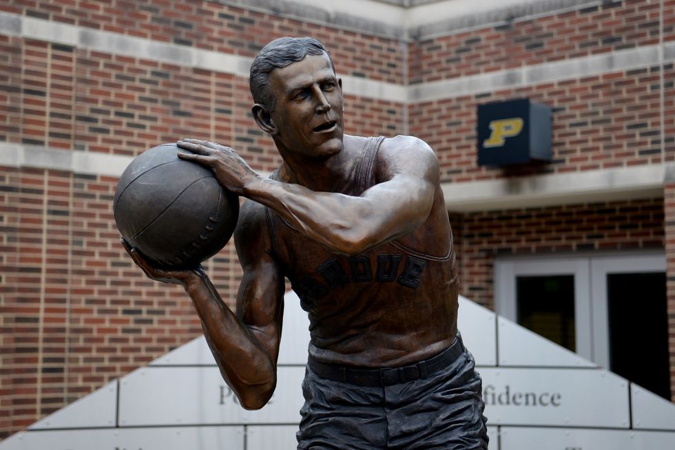 a bronze statue of john wooden wearing a basketball jersey and holding a basketball, located outside the mackey arena